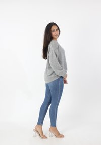 Grijze gebreide trui met een losse pasvorm, gecombineerd met blauwe skinny jeans en doorzichtige block-heeled sandalen. Witte achtergrond.