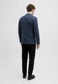 Tumes värviga struktuuriga bleiser, mille taga on vent, koos mustade pükste ja mustade kingadega, vaadates tagant.