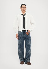 Camicia bianca a maniche lunghe con cravatta nera, abbinata a jeans a gamba larga blu con ricamo floreale bianco e un'etichetta marrone. Scarpe marroni.