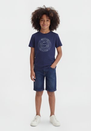 Enfant aux cheveux bouclés portant un t-shirt Moschino bleu marine, un short en jean et des baskets blanches, debout avec une main dans la poche.