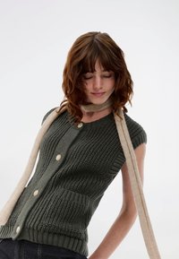 Καφέ πράσινο πλεκτό γιλέκο με κοντά μανίκια, υφαντό σχέδιο, κούμπωμα με κουμπιά και δύο τσέπες, συνδυασμένο με μπεζ κασκόλ.