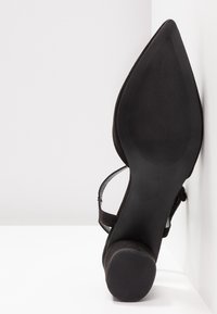 Semelle de chaussure noire à bout pointu, vue de face, avec une semelle texturée antidérapante et un talon rond, reposant contre une surface d'angle blanche.