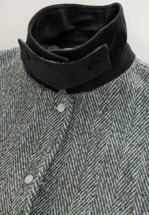 Grau gestreifte Stoffjacke im Fischgrätmuster mit einem schwarzen Cordkragen, ausgestattet mit Knopfverschlüssen und bunten Farbspritzern im Material.