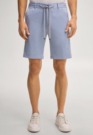 Mężczyzna ubrany w jasnoniebieskie teksturowane szorty ze sznurkiem w pasie oraz białe tenisówki, stojący na jednolitym tle.