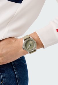 Kerek ezüstóra zöld számlappal, szürke részletekkel és rugalmas zöld pánttal, csuklón viselve, fehér hosszú ujjú felsővel kombinálva.