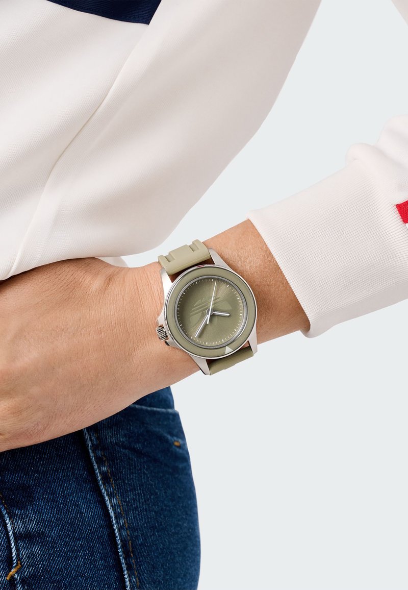 Kerek ezüstóra zöld számlappal, szürke részletekkel és rugalmas zöld pánttal, csuklón viselve, fehér hosszú ujjú felsővel kombinálva.