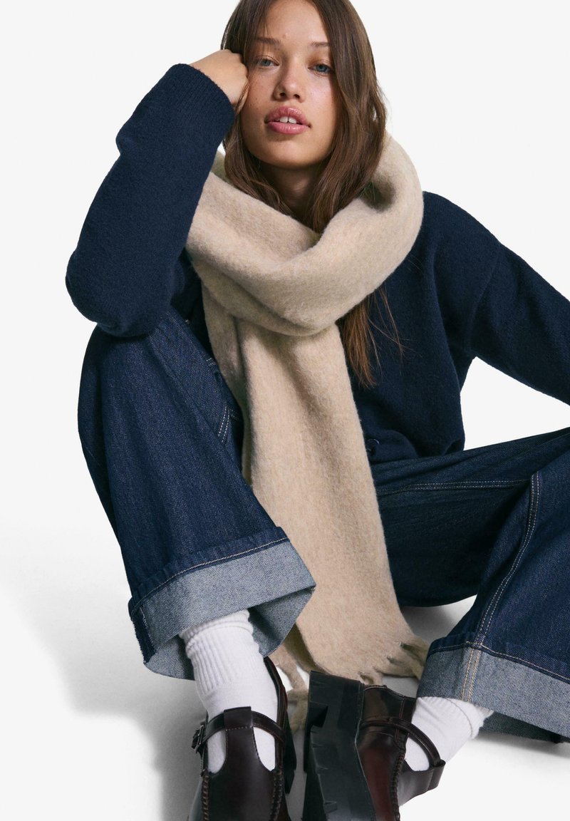 Écharpe en laine beige drapée sur un pull bleu marine, accompagnée de jeans en denim larges avec des poignets bleu clair, et des chaussures marron robustes.