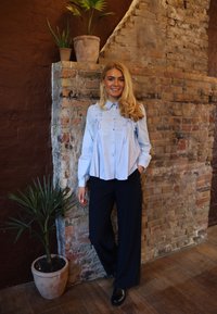 Gaiši zila krekla ar pogām un volānu detaļām, kopā ar plašām tumši ziliem biksēm. Fons ir redzams ķieģeļu siena un podos augs.