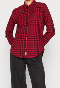Red and black plaid long-sleeve shirt with a collar, button closure, and a curved hem. Made from a textured fabric. Casual fit.