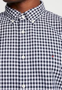 Chemise à carreaux bleu marine et blanc avec un col boutonné, dotée de boutons blancs et d'un petit accent de logo rouge, blanc et bleu.