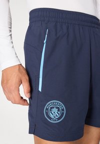Shorts de sport bleu marine avec des accents bleu clair, taille élastique, poche zippée sur le côté et logo brodé de Manchester City sur la jambe inférieure.