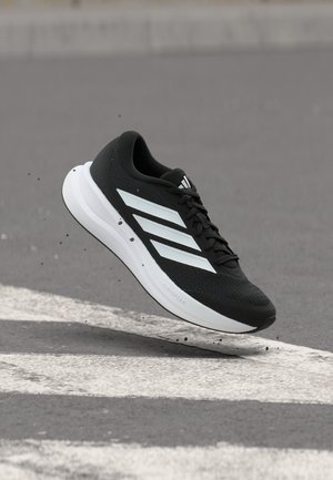 Schwarz-weißer Adidas Laufschuh mit weißen Streifen, schwebend über einer strukturierten Asphaltoberfläche mit weißen gemalten Linien.