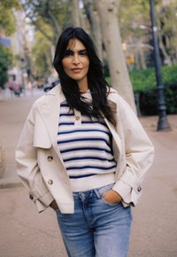Une femme aux longs cheveux bruns marche en plein air, portant une veste beige sur un pull à rayures bleues et blanches, ainsi qu'un jean bleu, les mains dans les poches.