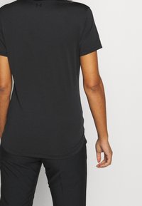 Personne portant un t-shirt noir ajusté à manches courtes Under Armour et un pantalon noir, vue de dos sur un fond uni.