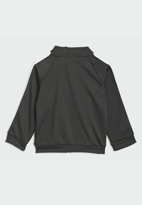 Veste de sport noire avec un col haut, des manches raglan et des bandes latérales blanches. Fabriquée en tissu lisse avec des poignets et un ourlet côtelés.
