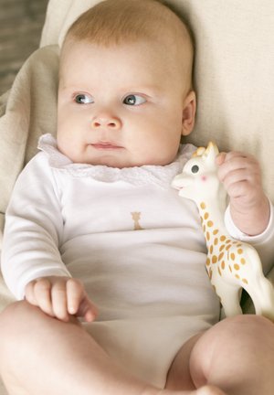 Neonato che indossa un body bianco, stringendo un giocattolo da dentizione a forma di giraffa, mentre guarda di lato mentre è seduto su un cuscino beige.