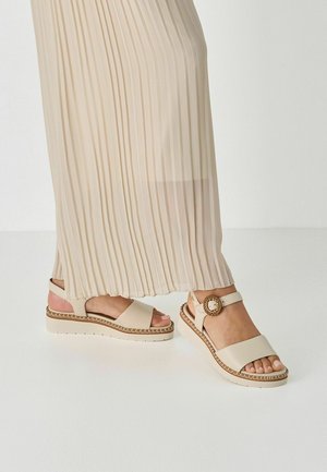 Beige platform sandals with a wide strap and ankle buckle. Textured sole with decorative stitching. Worn with a pleated light-colored skirt.