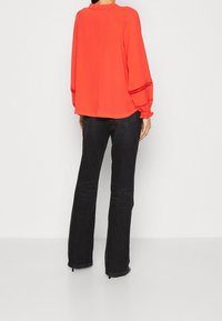 Personne portant un chemisier long à manches longues de couleur orange vif avec des poignets décoratifs et un jean noir taille haute, coupe droite, se tenant de dos.