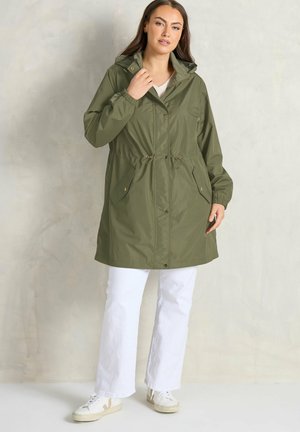 Mujer con impermeable verde oliva con capucha y cordones en la cintura, pantalones blancos y zapatillas blancas, de pie frente a una pared clara con textura.