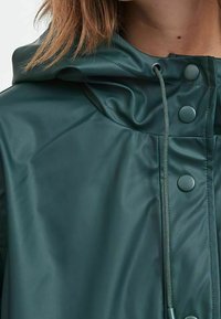 Mörkgrön vattentät jacka med en glänsande yta, med huva, dragsnöre och tryckknappar framtill för stängning.