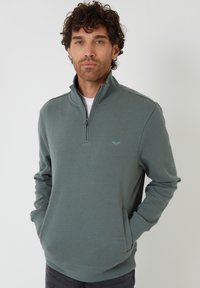 Zelený fleece pulovr s krátkým zipem, s jemnou strukturou, bočními kapsami a malým logem na hrudi. Dlouhé rukávy s žebrovanými manžetami.
