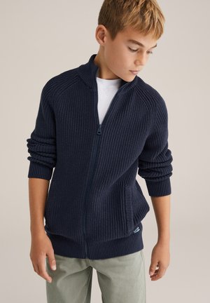 Navy modra zip-up jopica z rebrasto teksturo in zavitimi manšeti. Ima visok ovratnik in stransko žep, nosi se čez belo srajco.