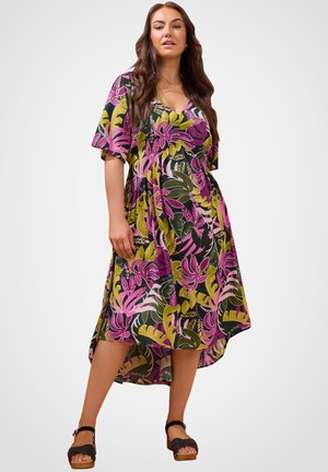 Mujer con vestido de estampado floral colorido con hojas en tonos rosa, verde y amarillo, combinado con sandalias negras de plataforma, posando sobre un fondo blanco.