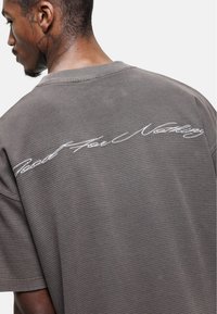 Grijze oversized T-shirt van een gestructureerde stof. Heeft cursieve witte tekst "Road For Nothing" op de achterkant. Vierkante vorm en losse pasvorm.