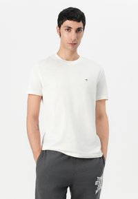Witte katoenen t-shirt met een ronde hals, korte mouwen en een klein logo op de borst. Het model draagt een grijze joggingbroek.
