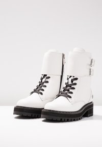 Botas de tobillo de cuero blanco con un diseño de caña alta, cordones negros, cremallera lateral y dos hebillas ajustables. Suela de goma negra gruesa.