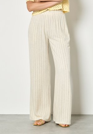 Femme portant un pantalon large crème à fines rayures verticales et des sandales à talons dorées à lanières, debout contre un mur uni.