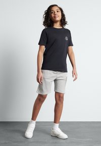 Svart t-skjorte med kort erme og liten logo på venstre bryst, kombinert med lysegrå shorts laget av mykt stoff, og hvite joggesko.