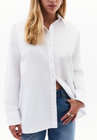 Camicia bianca con bottoni e maniche lunghe, realizzata in tessuto morbido, con colletto a punta e vestibilità rilassata. Abbinata a jeans in denim blu.