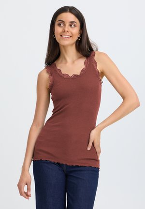 Mujer joven con cabello largo y oscuro lleva una camiseta marrón sin mangas con encaje y vaqueros azul oscuro, sonriendo y posando con una mano en la cadera.