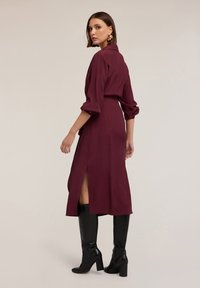 Frau in einem burgunderfarbenen, langärmligen Midikleid mit seitlichem Schlitz, trägt schwarze kniehohe Stiefel mit Absatz und goldene Creolen, schaut über die Schulter.