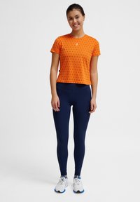 Oranje cropped t-shirt met kleine patronen, gecombineerd met marineblauwe legging en grijze sportschoenen met blauwe accenten. Gladde stoftextuur.