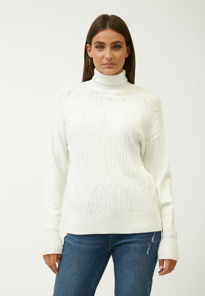 White knitted sweater with a turtleneck, featuring raised diamond patterns and ribbed cuffs. Worn with blue distressed jeans.