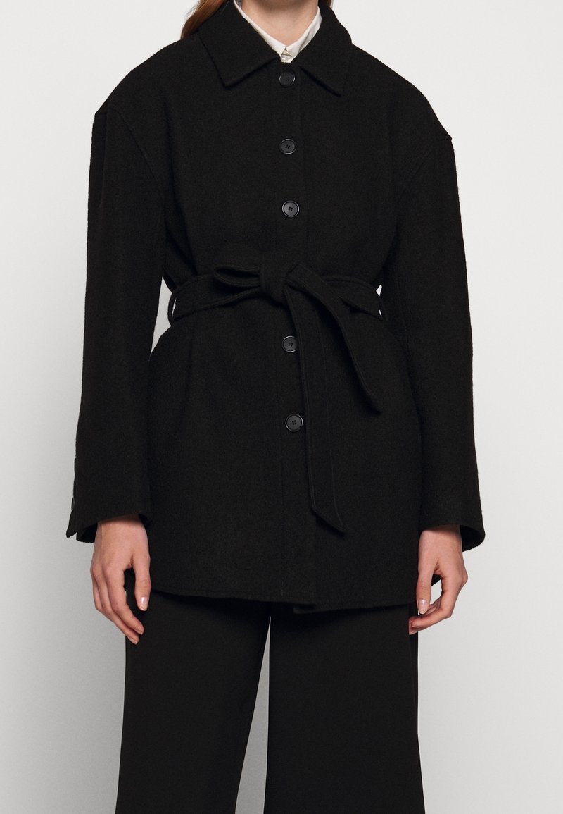 Manteau en laine noire avec une fermeture boutonnée sur le devant, une taille ceinturée et des manches longues. Comprend un col et de gros boutons. Tissu texturé.