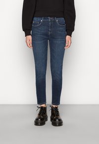 Jeans skinny azul-escuro feitos de denim, com uma cintura de altura média, cinco bolsos e desbotamento subtil. Combinados com botas pretas chunky.