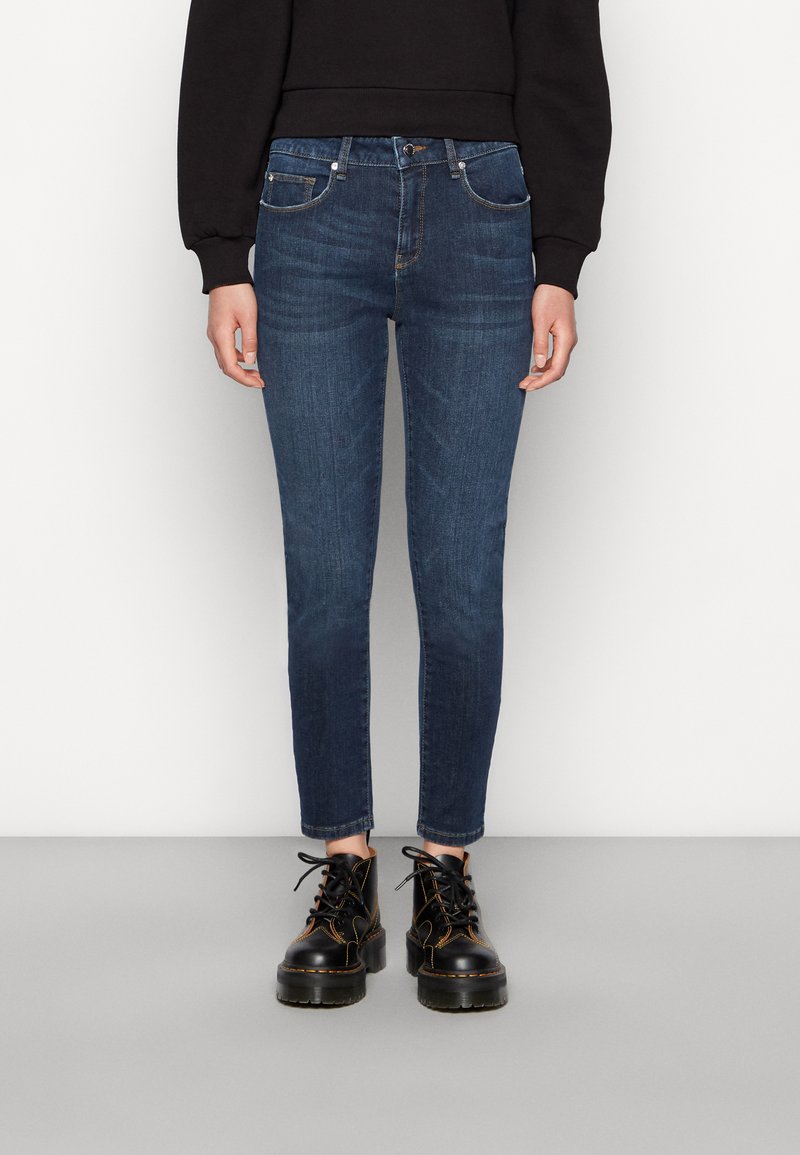 Jeans skinny azul-escuro feitos de denim, com uma cintura de altura média, cinco bolsos e desbotamento subtil. Combinados com botas pretas chunky.