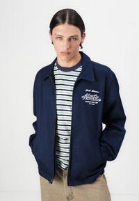 Navy blue zippered jacket with a pointed collar, paired with a green and white striped t-shirt. Features logo detailing on the chest.