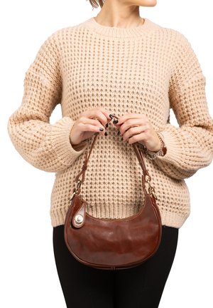 Borsa a tracolla in pelle marrone con forma arrotondata, dotata di una tracolla corta e un accento decorativo. Indossata con un maglione beige chunky e leggings neri.
