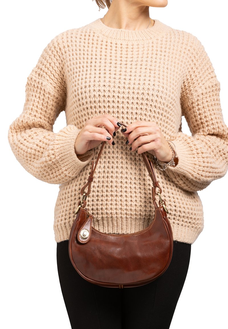 Borsa a tracolla in pelle marrone con forma arrotondata, dotata di una tracolla corta e un accento decorativo. Indossata con un maglione beige chunky e leggings neri.