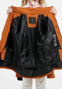 Orange Jacke mit schwarzem Innenfutter, ausgestattet mit einem Etikett, mehreren Taschen und einer glatten Textur. Auffällige Details sind kontrastierende Einsätze und Beschläge.