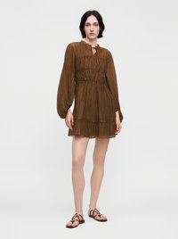 Jeune femme aux cheveux courts et foncés, portant une robe plissée marron à manches longues et des sandales noires, debout devant un fond clair uni.