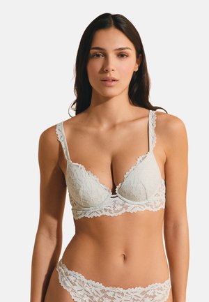 Kobieta w białym koronkowym biustonoszu i dopasowanej koronkowej bieliźnie, stojąca i zwrócona przodem na jednolitym tle.