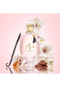 Flacon de parfum Juicy Couture Just Moi avec un bouchon doré, entouré de fleurs blanches, de gousse de vanille, de morceaux de chocolat et d'ambre sur un fond rose.