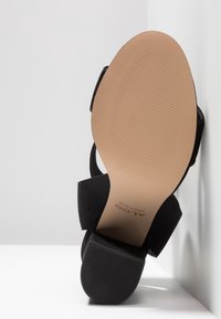 Sandale noire à talon avec un dessus texturé et une semelle lisse. La semelle présente des rainures horizontales pour une meilleure adhérence et un léger relèvement du talon.