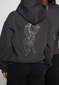 Dunkelgrauer Hoodie mit einem großen, mehrfarbig bestickten Motiv auf dem Rücken, das eine Figur zeigt, die mit floralen Elementen verflochten ist.