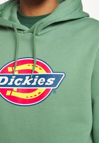 Sudadera verde con un gran logotipo gráfico "Dickies" en azul y rojo con acentos amarillos, hecha de una tela suave y texturizada.